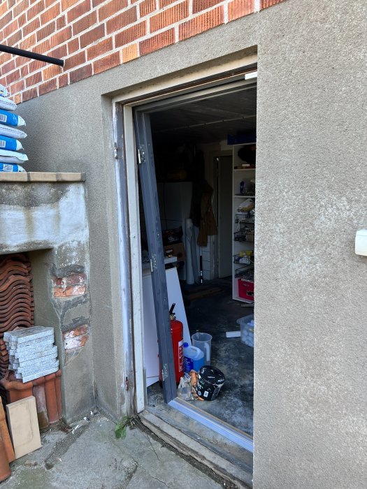 Öppen dörr till en renoveringsplats med synliga byggmaterial och verktyg. En oorganiserad arbetsyta med förvaringshyllor i bakgrunden.