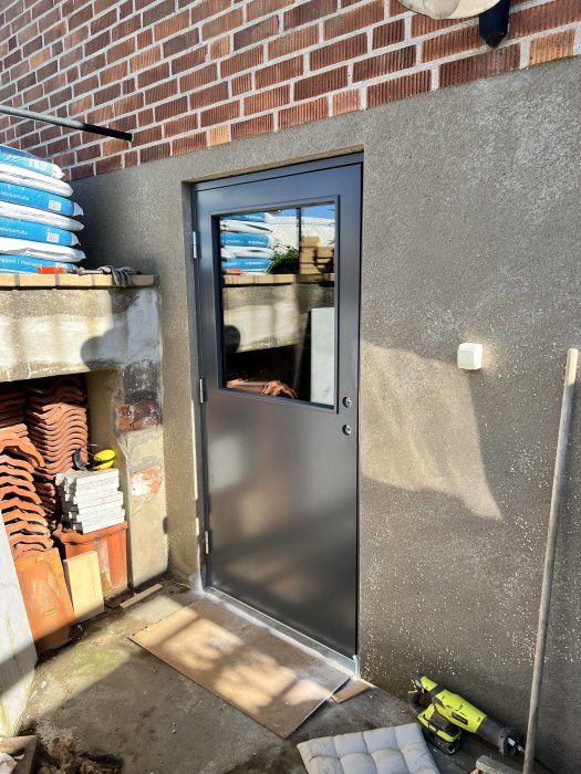 En modern, grå dörr med fönster, placerad i en vägg av puts och omgiven av byggmaterial och tegel.