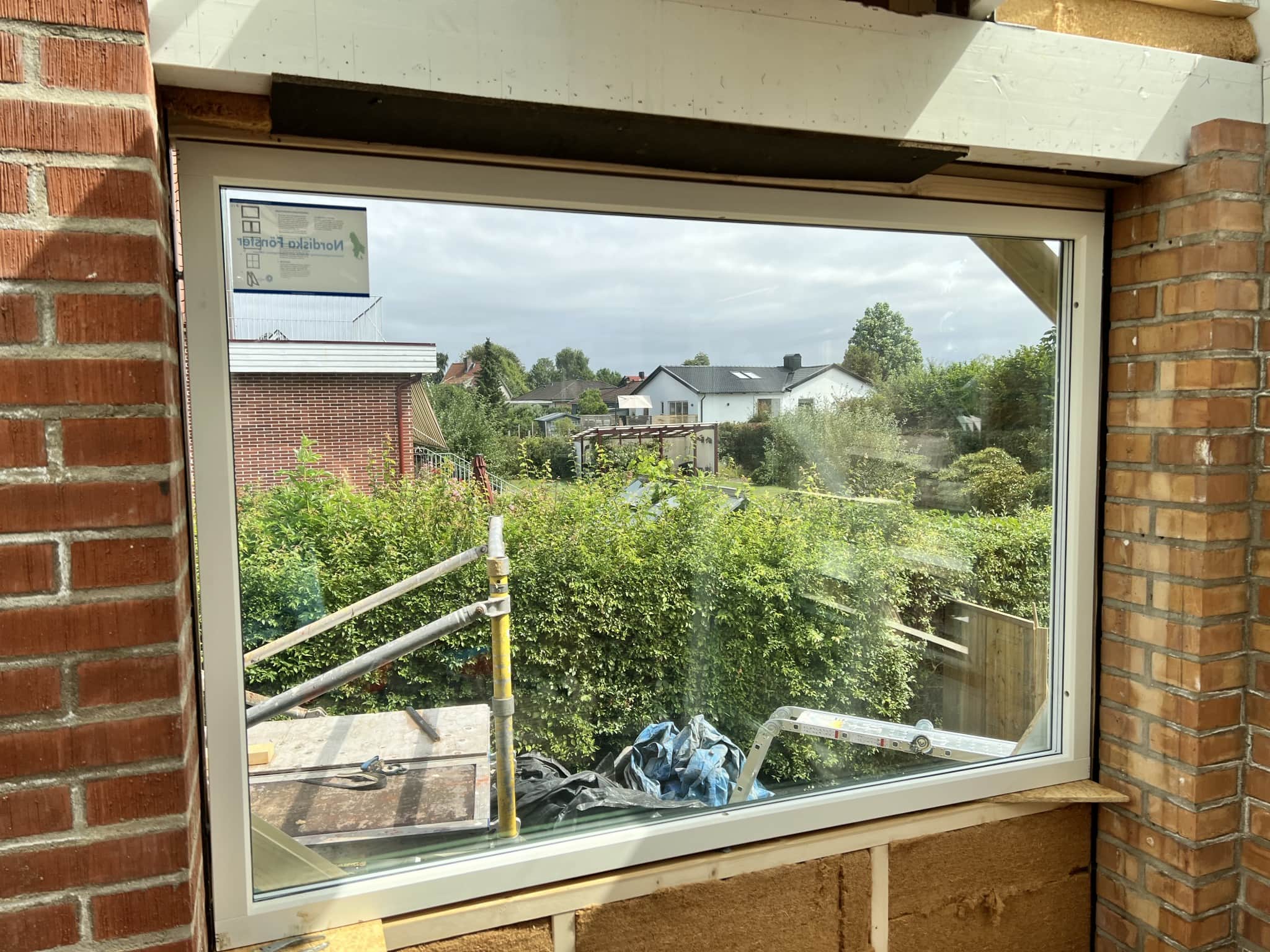 En stor, modern fönsteröppning med utsikt över en grönskande trädgård och närliggande hus, under renovering.