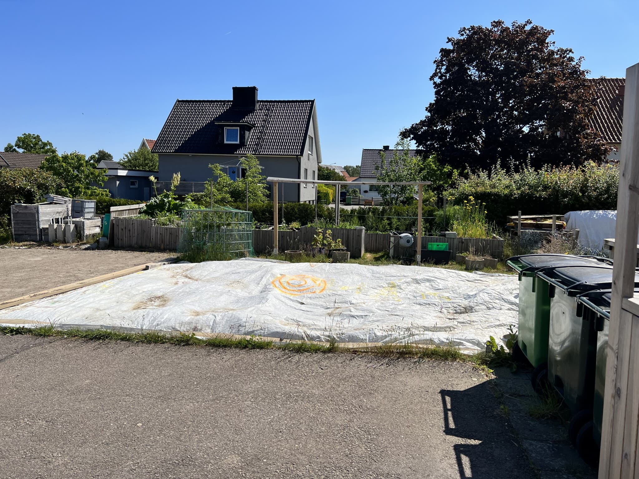 En solig tomt med en plastduk, omgiven av grönska och avfallskärl, i en lugn bostadsområde. Perfekt för framtida renoveringsprojekt.