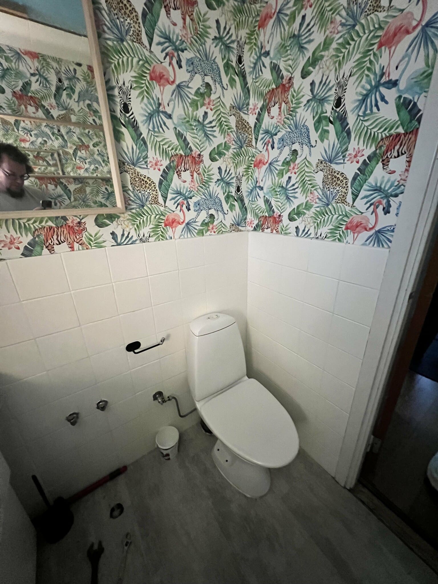 Färgglatt badrum med djungeltapet, vita kakelplattor och modern toalett. Verktyg på golvet antyder pågående renovering.