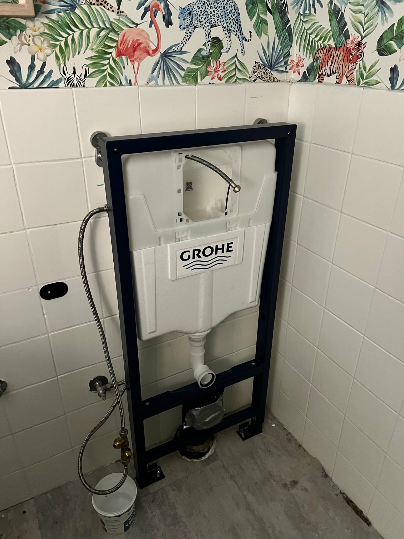 Väggmonterad toalettinstallation med Grohe-cistern, omgiven av tropiskt mönstrad tapet och vita kakelplattor. Perfekt för en uppfriskande badrumsrenovering.