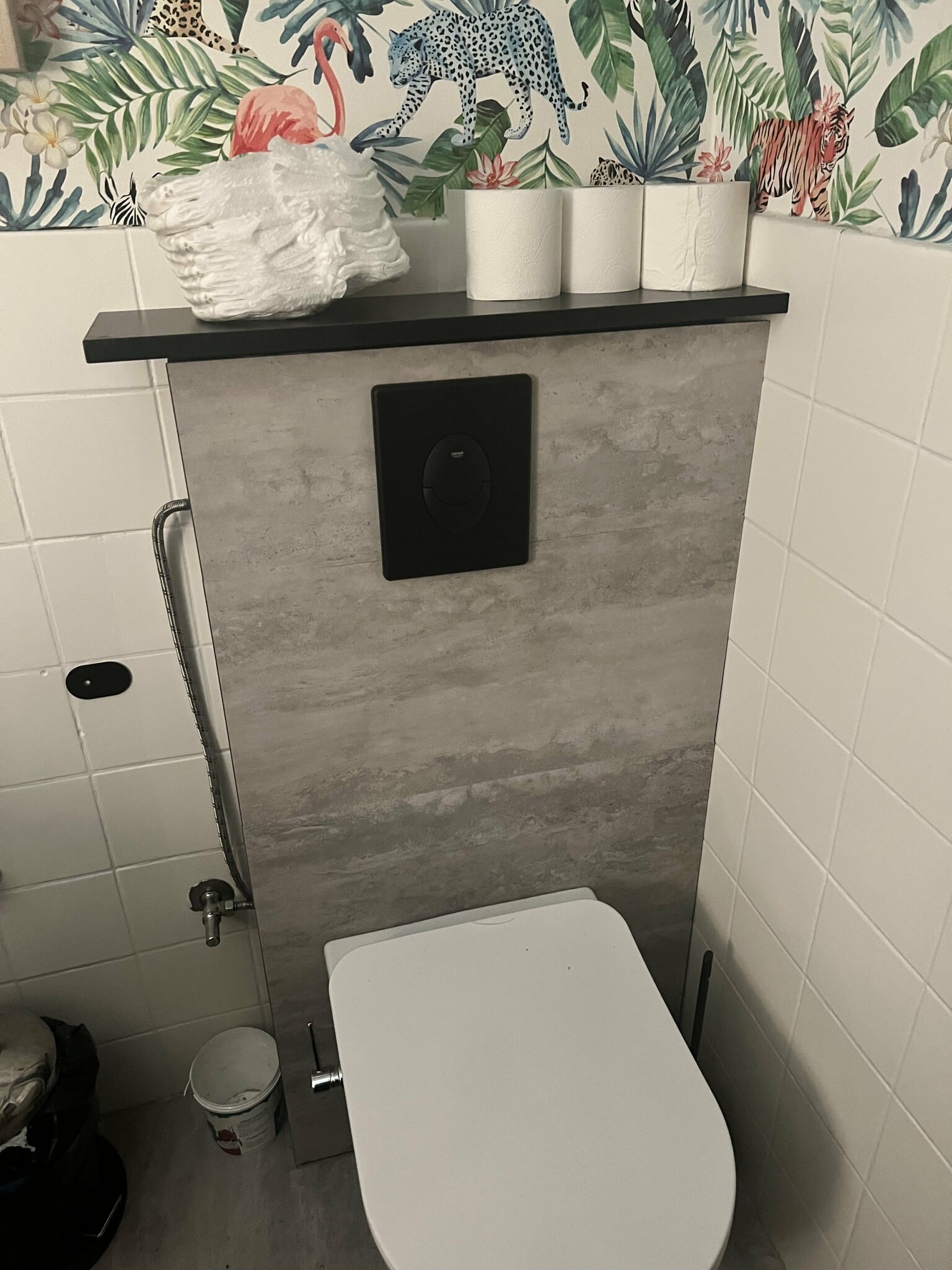 Modernt badrum med djungeltapet, väggmonterad toalett och dekorativ hylla med toalettpapper. Stilfull kontrast mellan betong och färgglada mönster.