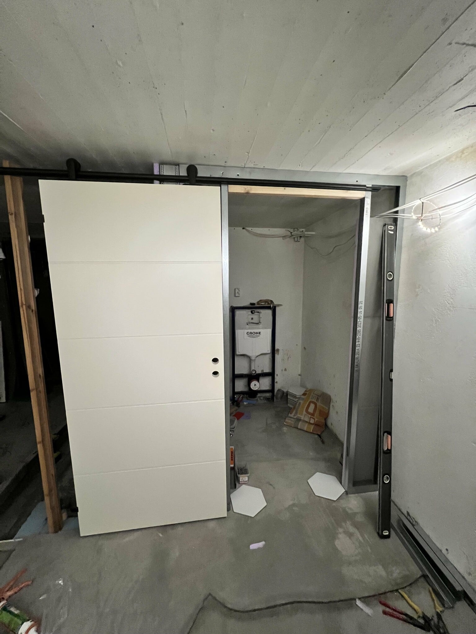 Källarrenovering med en skjutdörr av trä och synlig rördragning. Grohe enhet i bakgrunden. Verktyg och plattor på golvet.