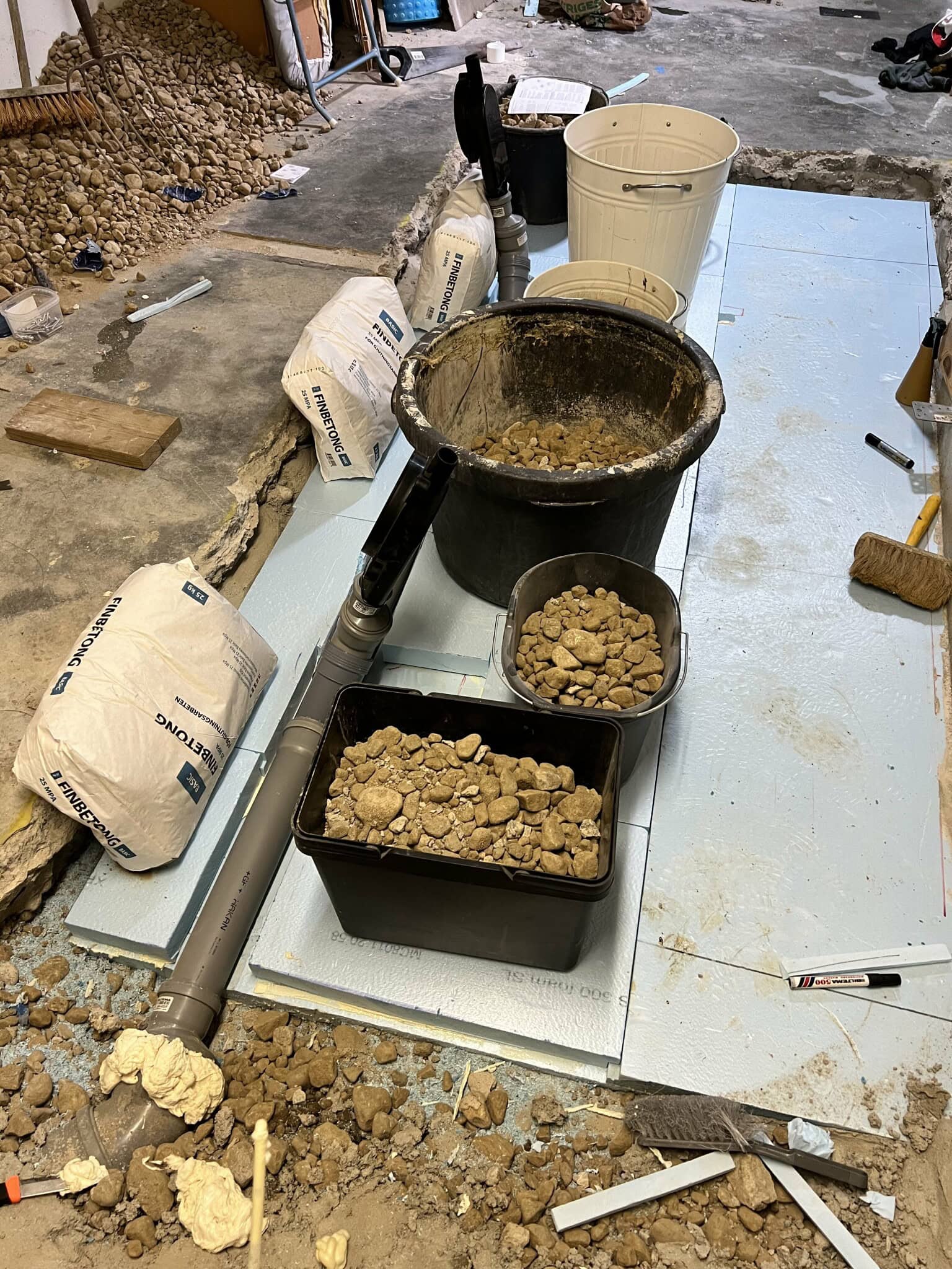 Renoveringsprojekt pågår: Verktyg, hinkar och säckar med fyllnadsmaterial på en byggarbetsplats, redo för golvläggning och dränering.