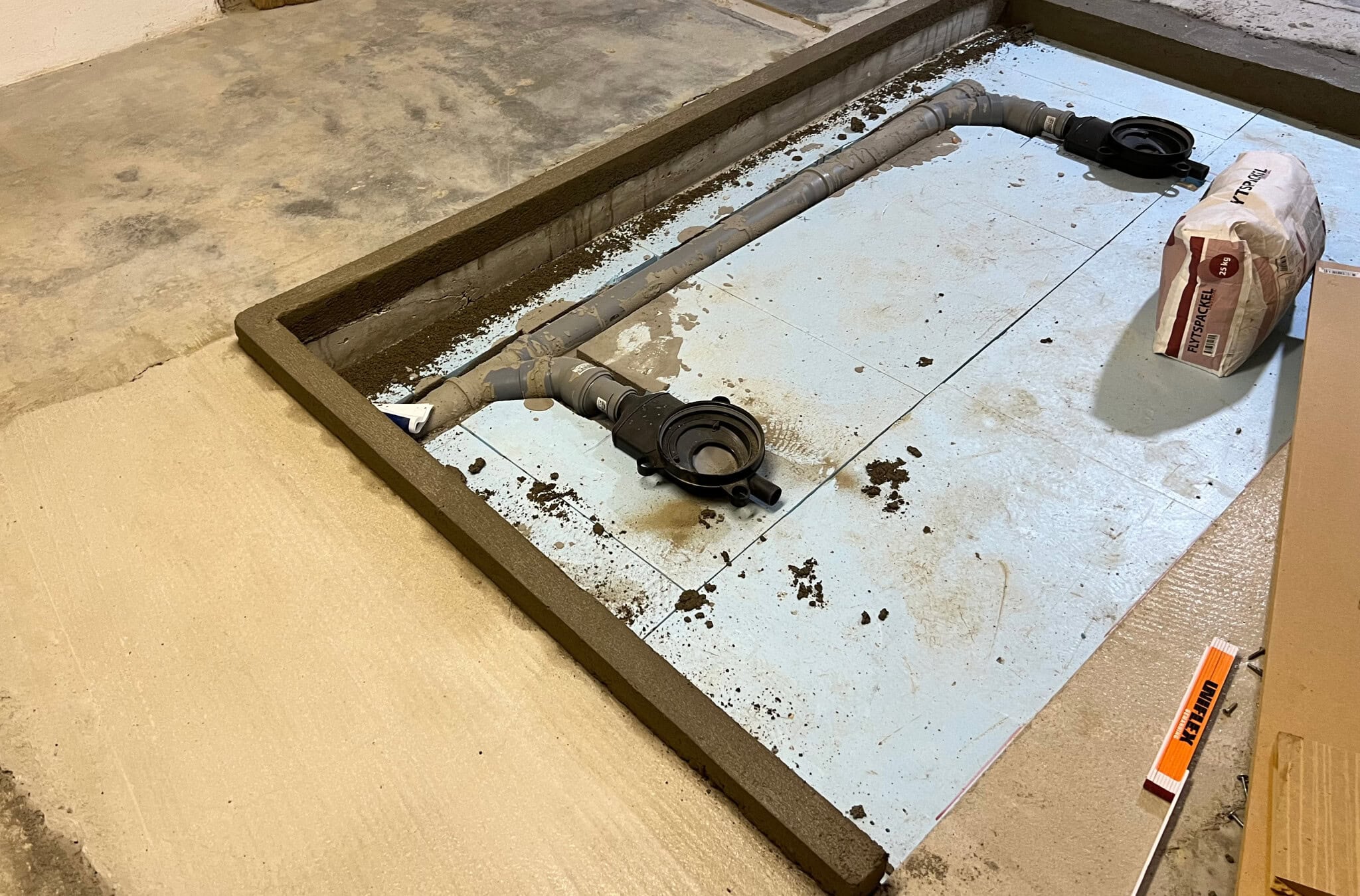 Vattenrörsinstallation i ett badrumsprojekt, med betongram och byggmaterial förberett för golvbeläggning. Perfekt för DIY-renovering.
