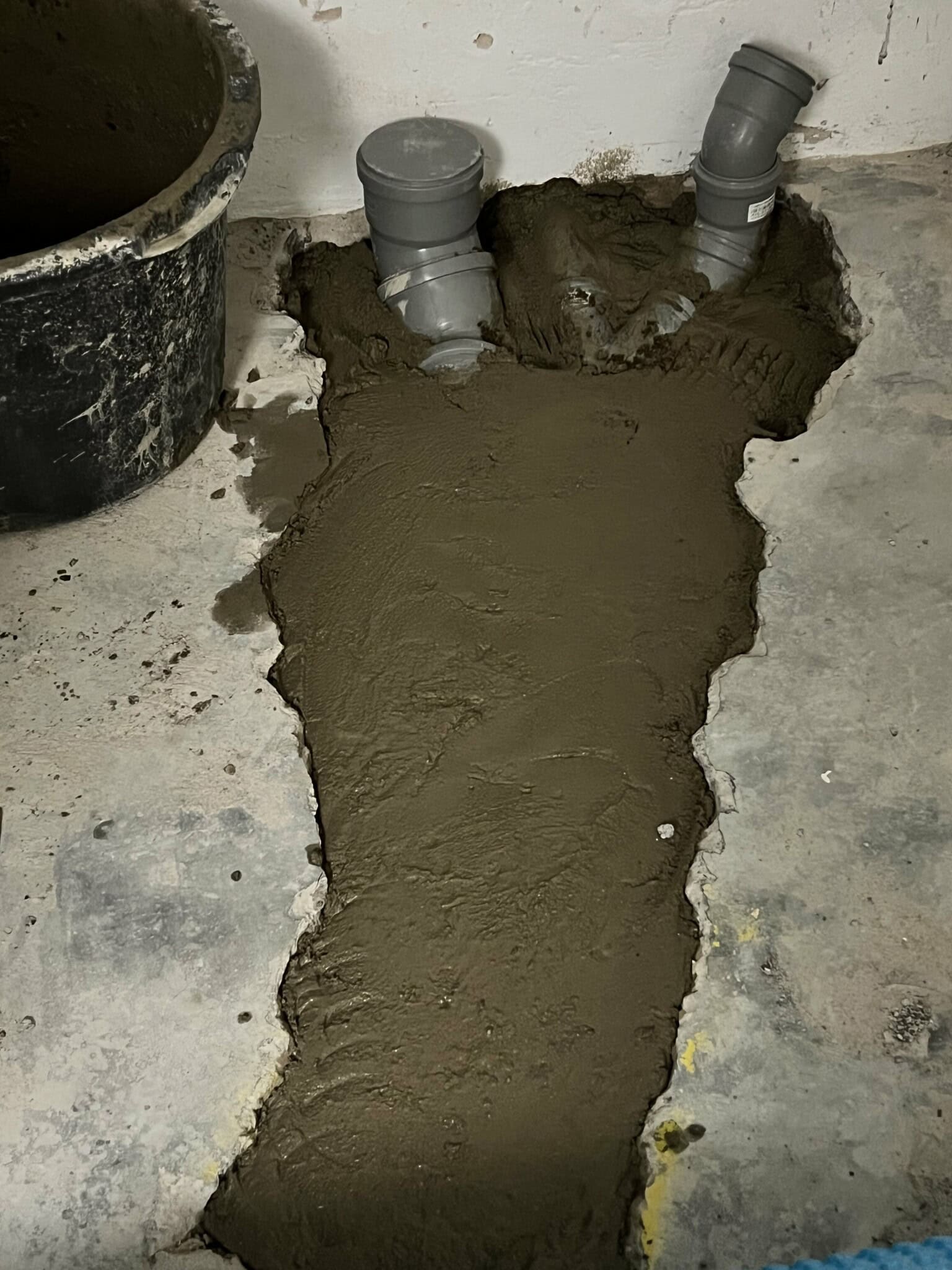 Nyinstallerade rör i ett betonggolv, omgivna av nylagd cementblandning. En hink står bredvid på det ojämna golvet.