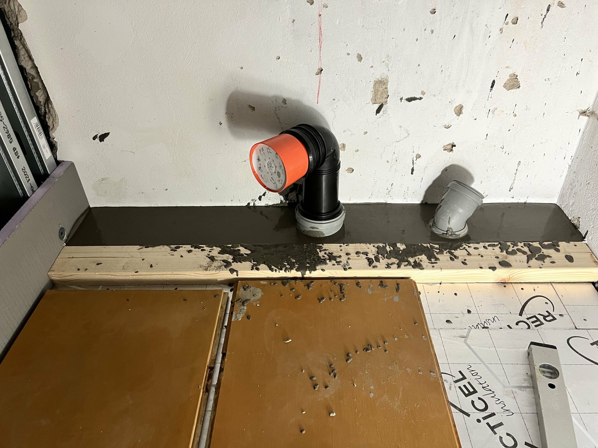 Renoveringsprojekt med installation av avloppsrör i betongvägg. Synliga byggmaterial inkluderar trä, betong och rördelar. Arbetsplatsen visar pågående arbete.