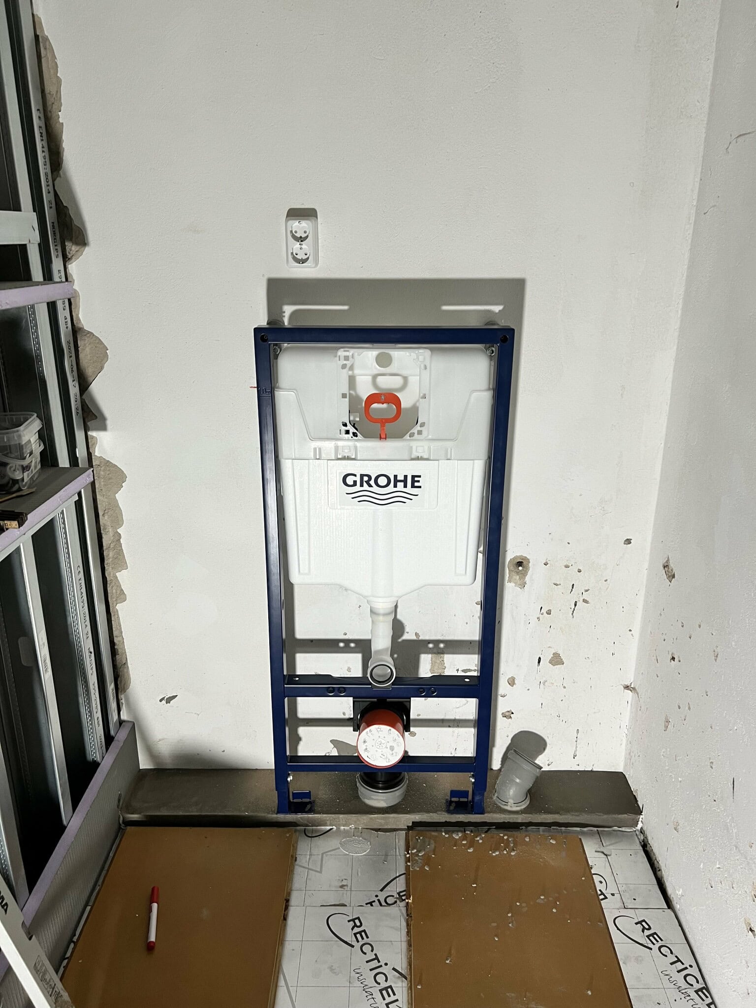 Väggmonterad toalettinstallation med Grohe-cistern under renovering. Omgiven av byggmaterial och verktyg. Förberedelse för kakelsättning och rördragning.