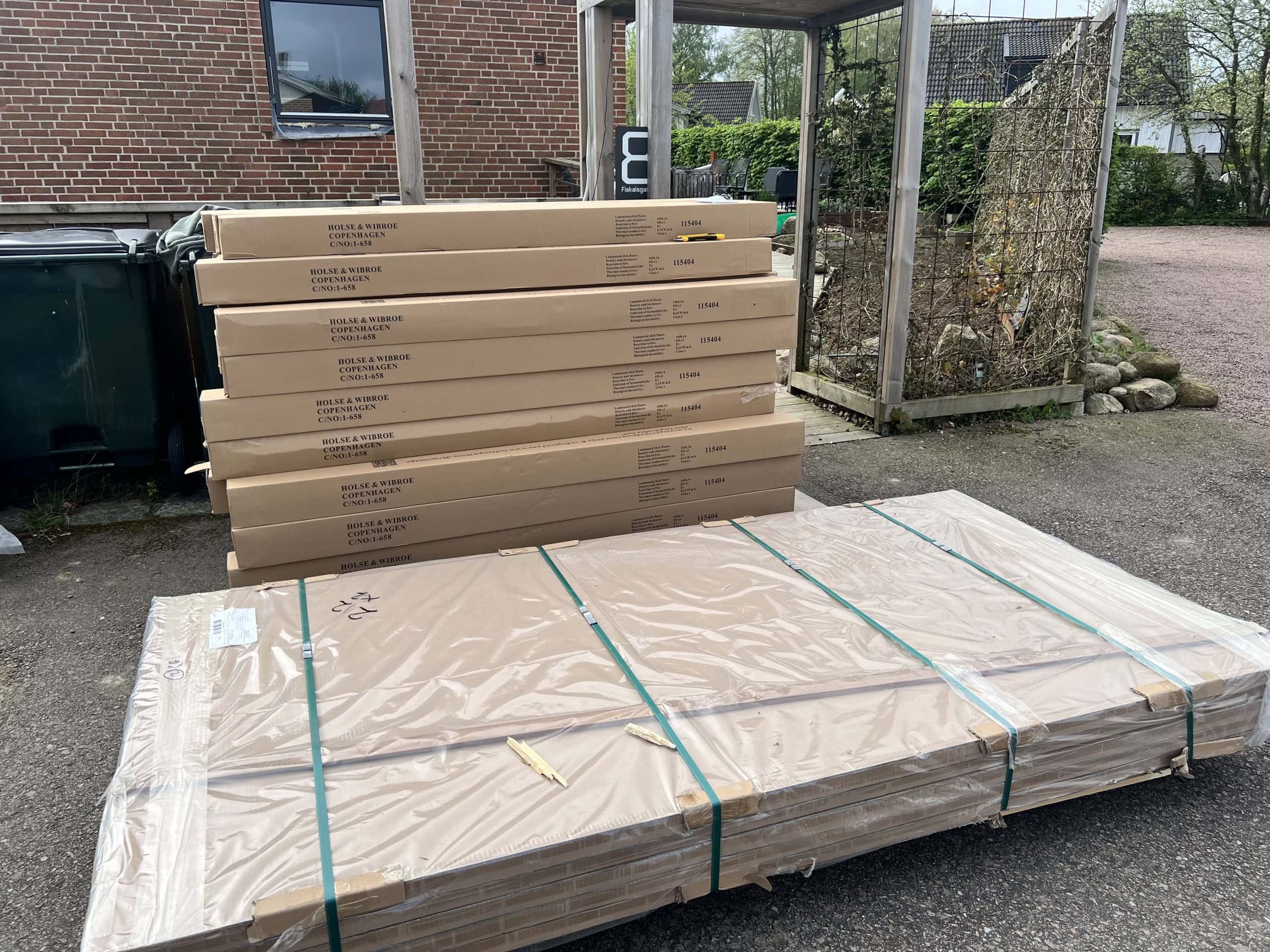 En hög med flera lådor och paket ligger staplade utomhus på marken vid en tegelbyggnad. Lådorna är märkta med "House & Wibroe Copenhagen". Det finns även en större, platt förpackning inslagen i plast och säkrad med band.